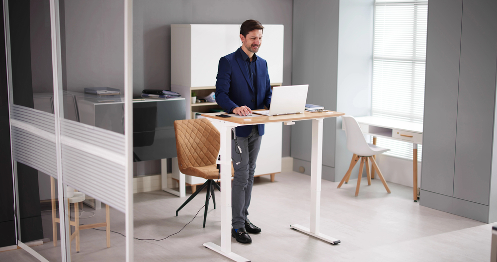 A man in a professional office setting using an adjustable standing desk while working on a laptop. The office features modern furniture, ample natural light, and a minimalist design.